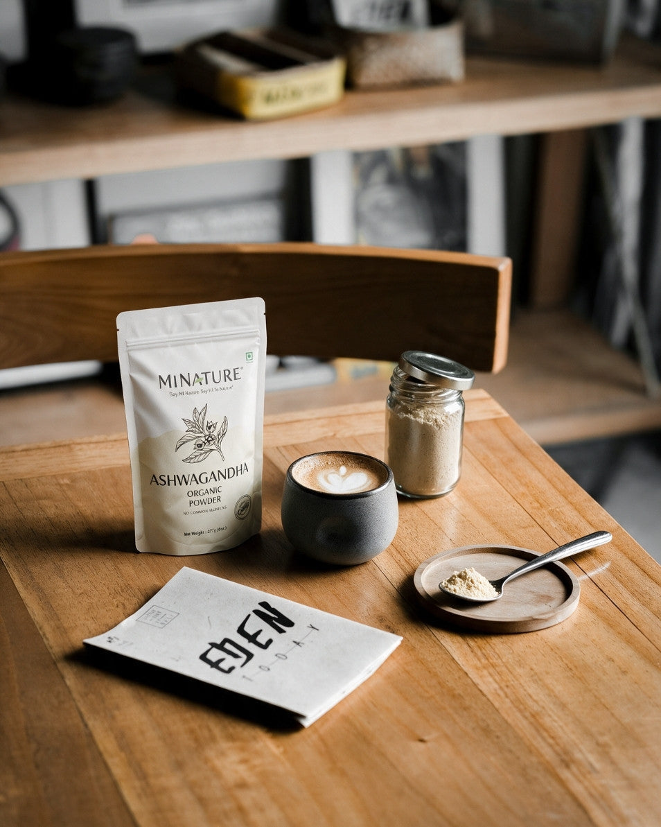 A styled tabletop scene showing a white stand-up pouch labeled MINATURE Ashwagandha Root Extract powder, a small dark ceramic cup with latte art, a glass jar of powder, a spoon on a small dish, and a folded paper card in print, all arranged on a wooden table in a warm indoor setting.
