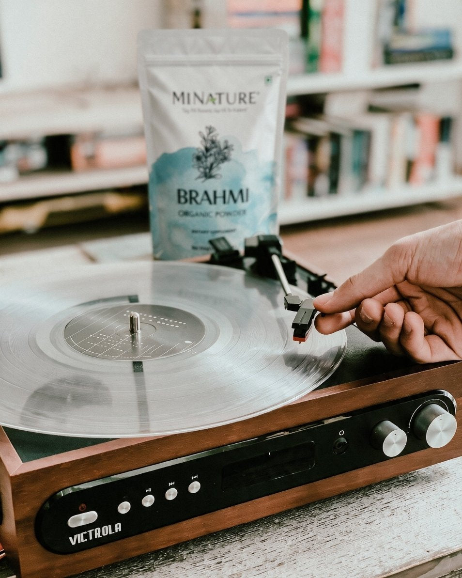 A hand is placing a record needle onto a vinyl record on a brown wooden Victrola turntable. A standee of Minature Brahmi Organic Powder is positioned behind the turntable, with a softly blurred bookshelf in the background.