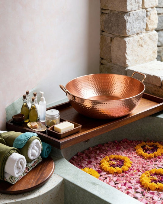 Copper bowl on a wooden tray with bath items next to a stone wall and bathtub with flowers.