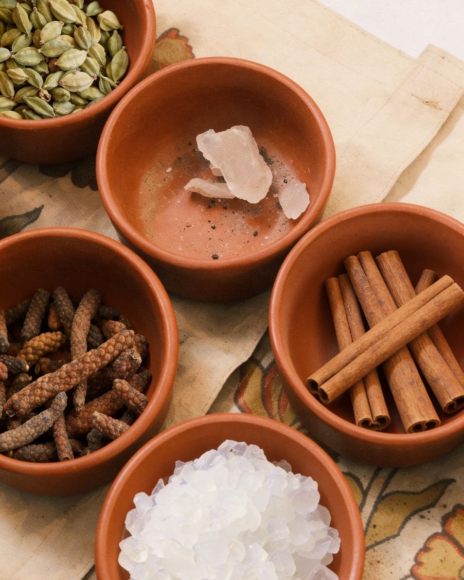 Five terracotta bowls containing various spices and ingredients on a patterned fabric background.